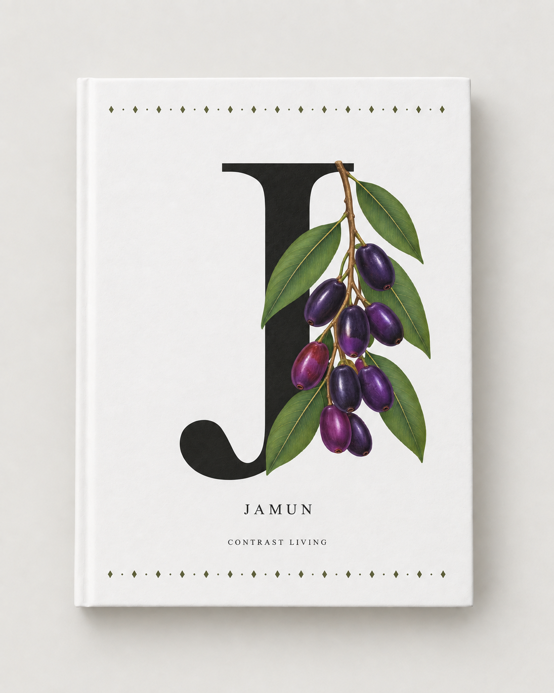 A hardcover journal or notebook with a white cover, featuring a large black letter 'J' overlaid with a botanical illustration of green leaves and purple jamun berries. The word 'JAMUN' is printed below, with 'CONTRAST LIVING' underneath, on a light gray background.