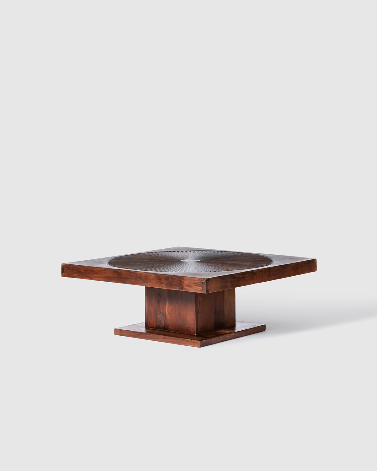 A square wooden coffee table with a dark metal circular inlay on the tabletop and a block-style pedestal base