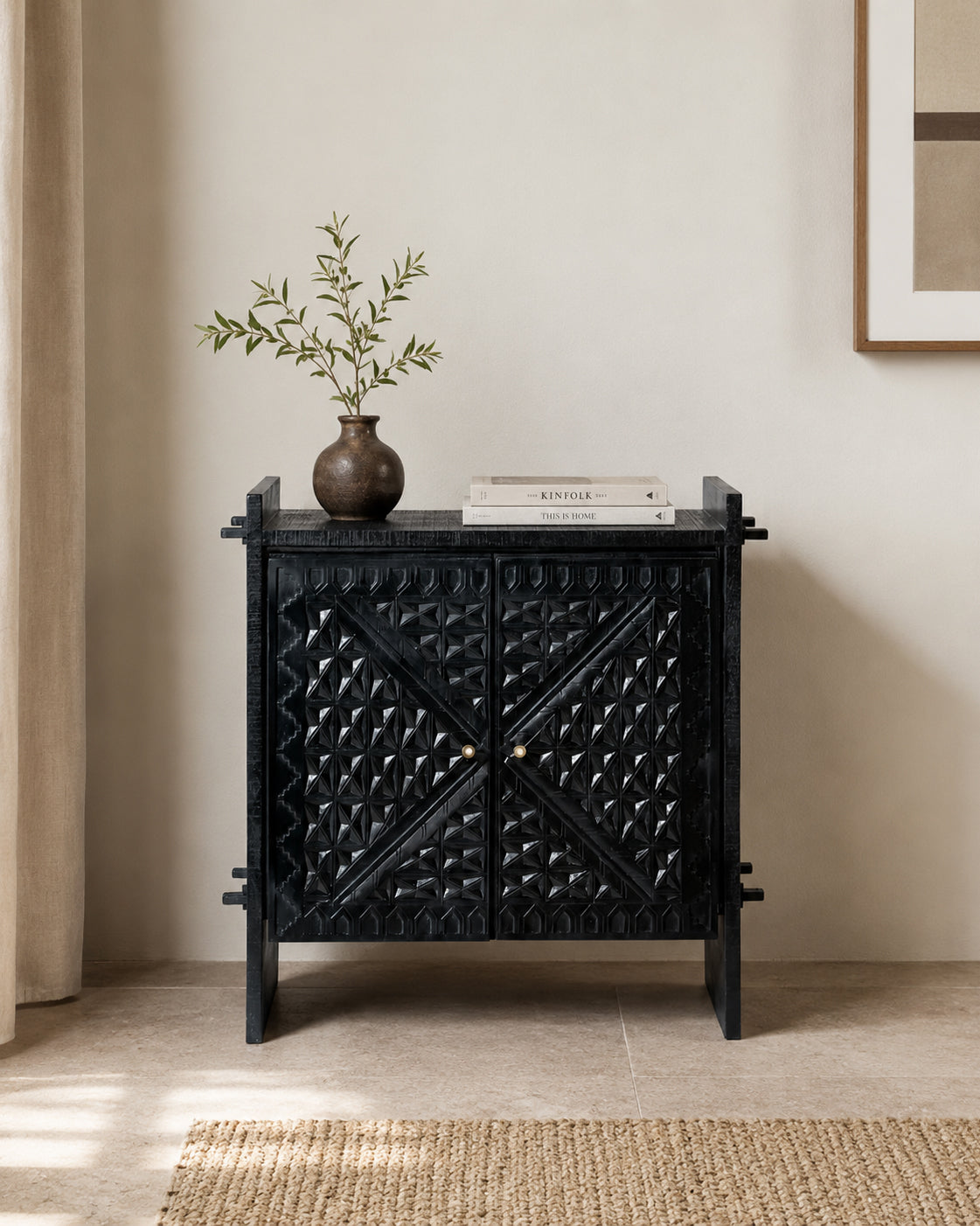 A black carved wooden cabinet with decorative geometric patterns on the doors, styled with a small vase of greenery and books on top, set against a neutral beige wall and floor.