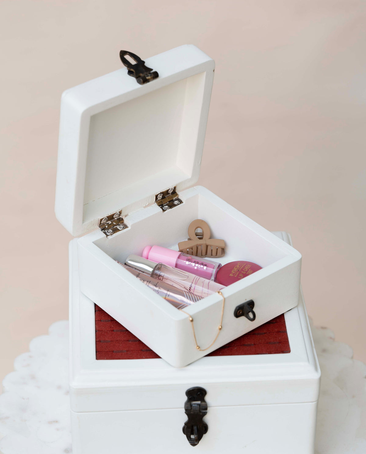 A white wooden jewelry box with black metal latches, shown open to display cosmetics inside, including a pink lip gloss, a clear tube, and a small wooden hair claw clip.