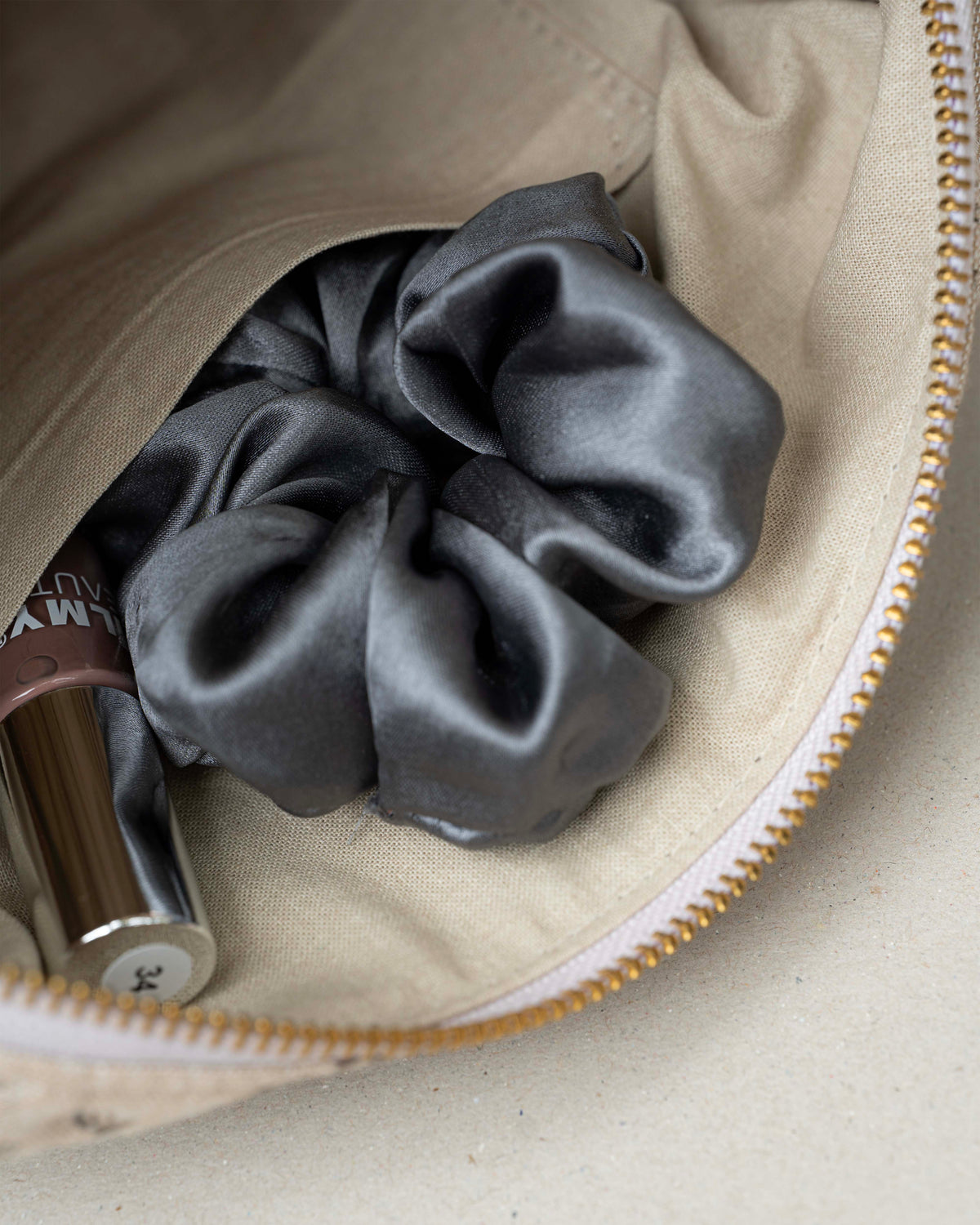A dark gray satin scrunchie nestled inside a beige fabric zippered pouch