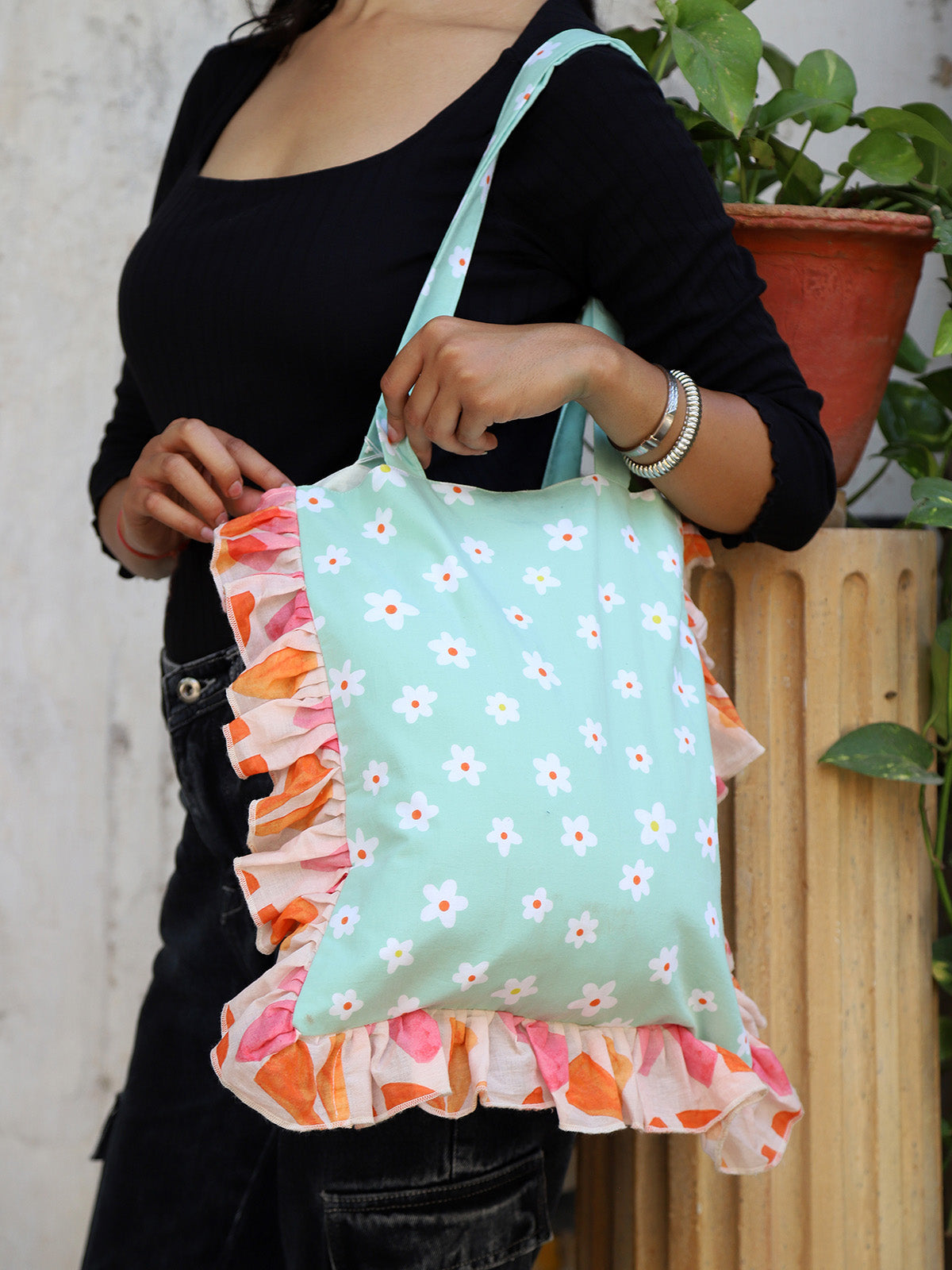 A person holding a tote bag with a mint green base, decorated with a white floral print and a vibrant, eye-catching ruffle border in pink and orange.