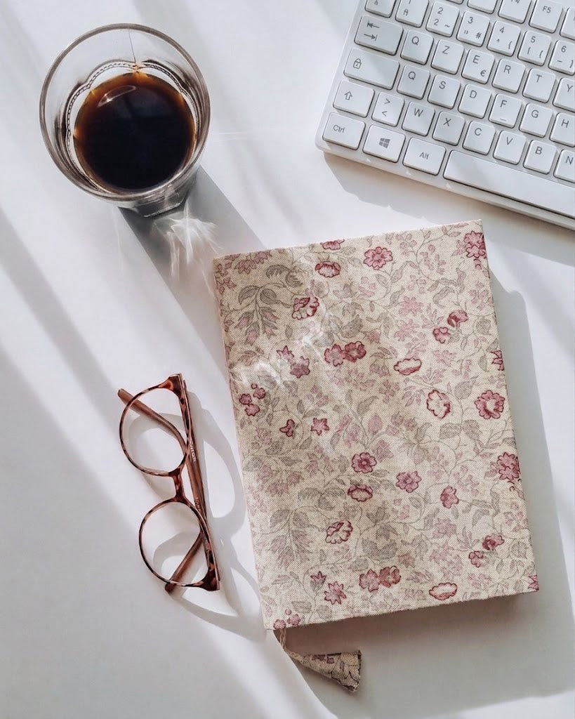 Floral notebook, glasses, and coffee on a white surface with a keyboard in the background
