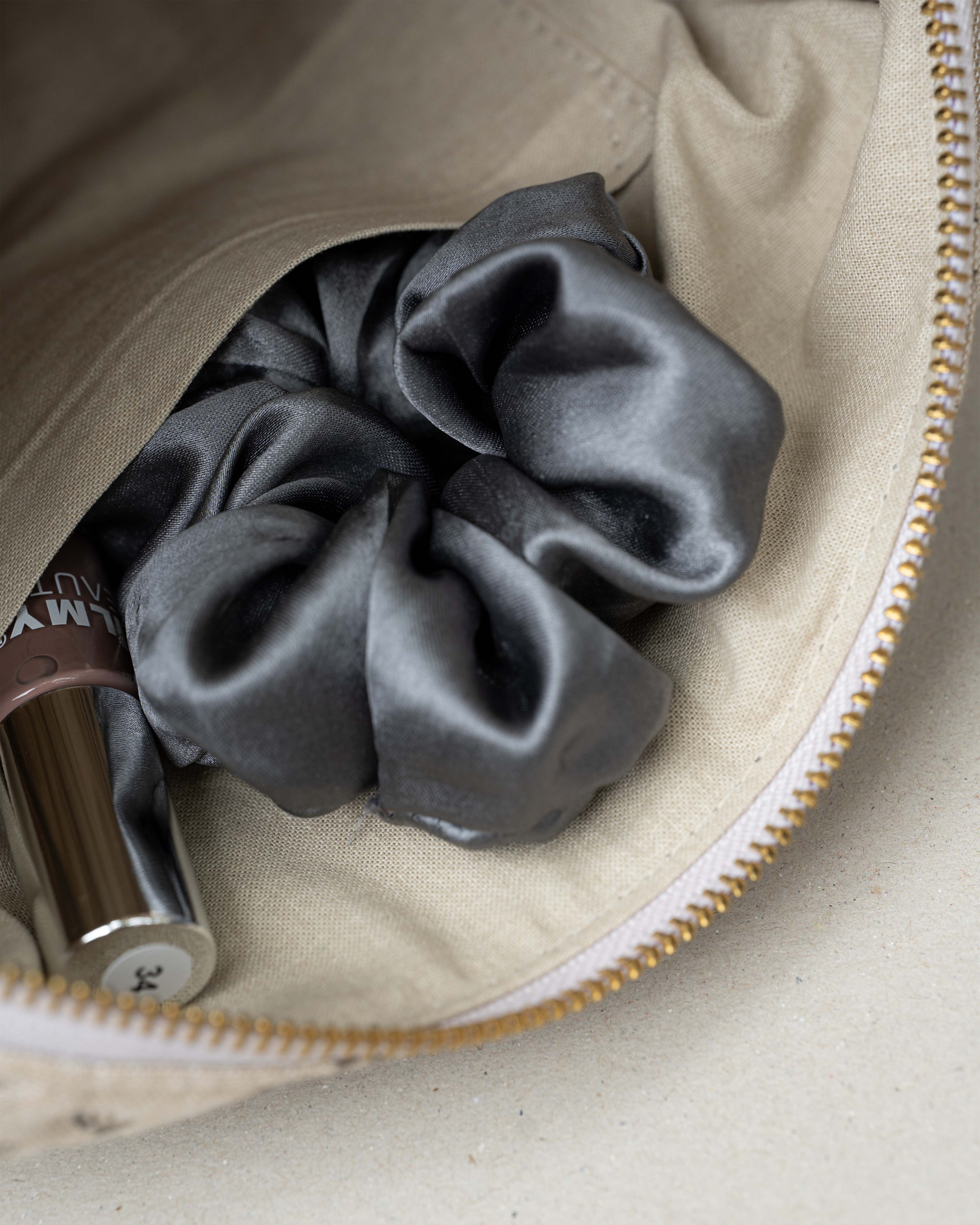 A dark gray satin scrunchie nestled inside a beige fabric zippered pouch