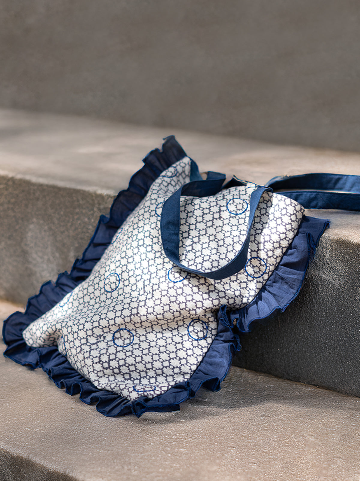 A handcrafted cotton tote bag with an indigo block-printed base and hand-embroidered circular motifs, complemented by sheer navy ruffle trim and long shoulder straps.
