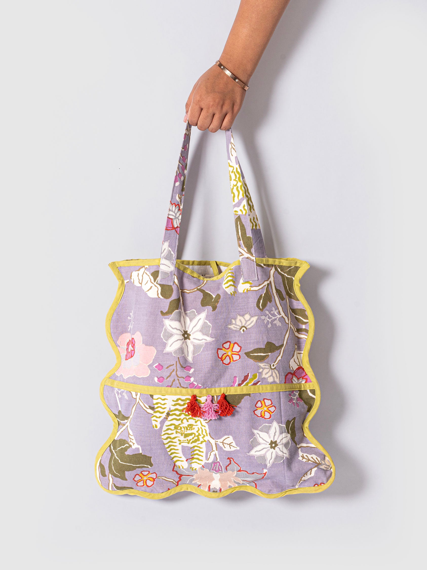 A handcrafted scallop-edged tote bag with a lavender-hued base featuring a bold hand-printed design with tigers, flowers, and foliage, finished with lemon-yellow piping and red tassels on the front pocket.