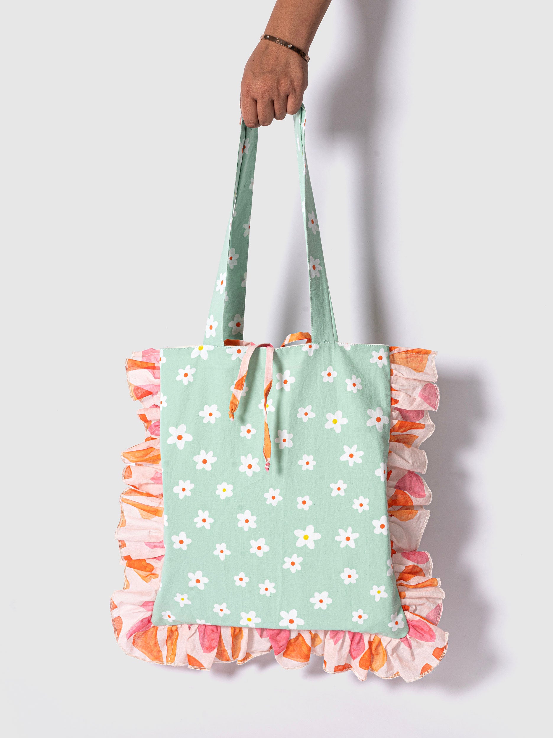 A tote bag with a mint green base covered with a white floral print, and a vibrant ruffle border in shades of pink and orange. 
